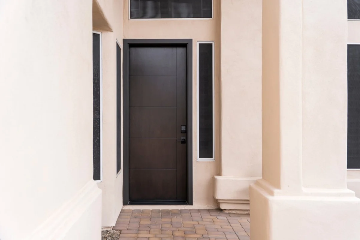 An inscribed, 8-foot Signet Cherry front entry door from ProVia stained in a rich Coffee Bean finish, framed by narrow vertical sidelites and a transom window. The dark woodgrain contrasts with the smooth, light stucco exterior and stone paver entryway.