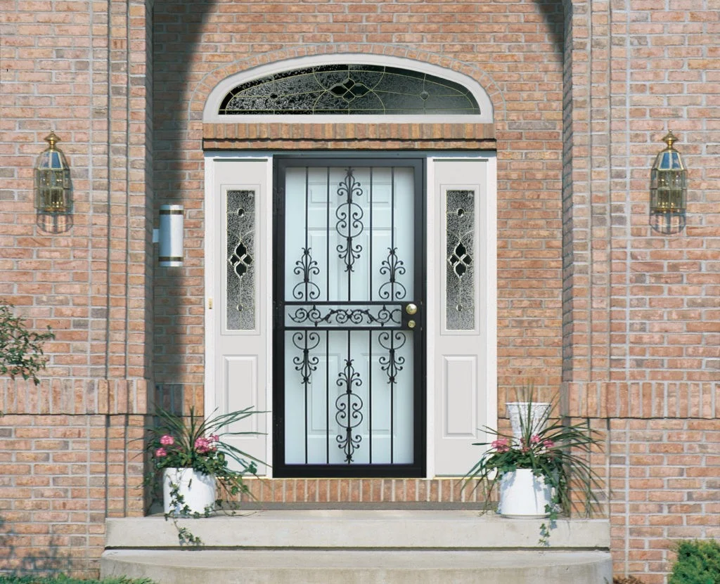 Security Door Installation | Ny Metro, Tri-State | Window King A stylish residential entryway featuring a white front door framed by decorative glass sidelights and a matching arched transom window. A black wrought iron security door from Home Guard with an ornate scroll design provides both protection and elegance. The home’s exterior is finished with light red brick, complemented by brass wall lanterns on either side of the doorway and potted plants flanking the steps.