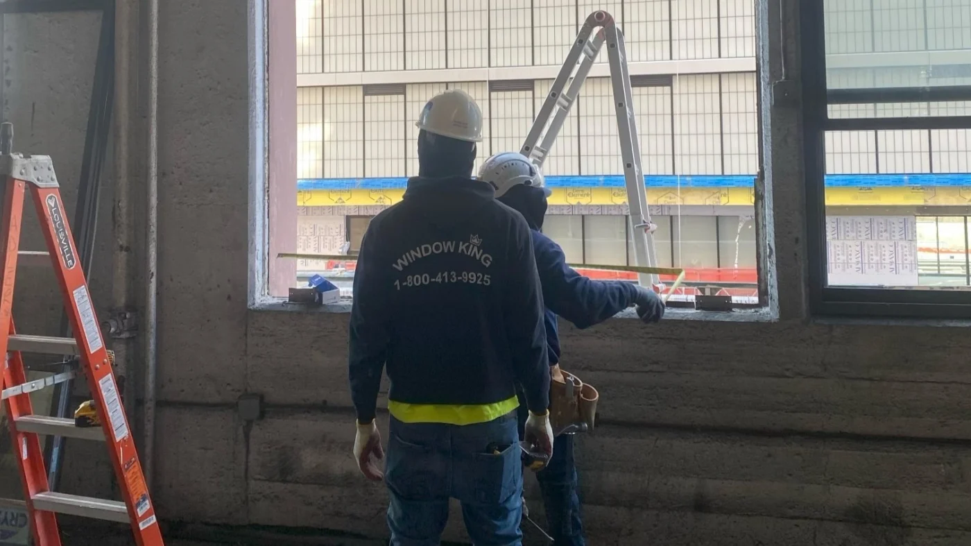 Inside Our Process | NY Metro Window Installation & Door Replacement | Window King Two Window King crew members on a job site, preparing a large opening for a new window installation. Both workers are wearing hard hats and safety gear, with one in a Window King jacket clearly displaying the company’s name and phone number. A bright orange ladder and tools are visible nearby, while natural light streams in through the open window frame, highlighting the precision and professionalism of their work.
