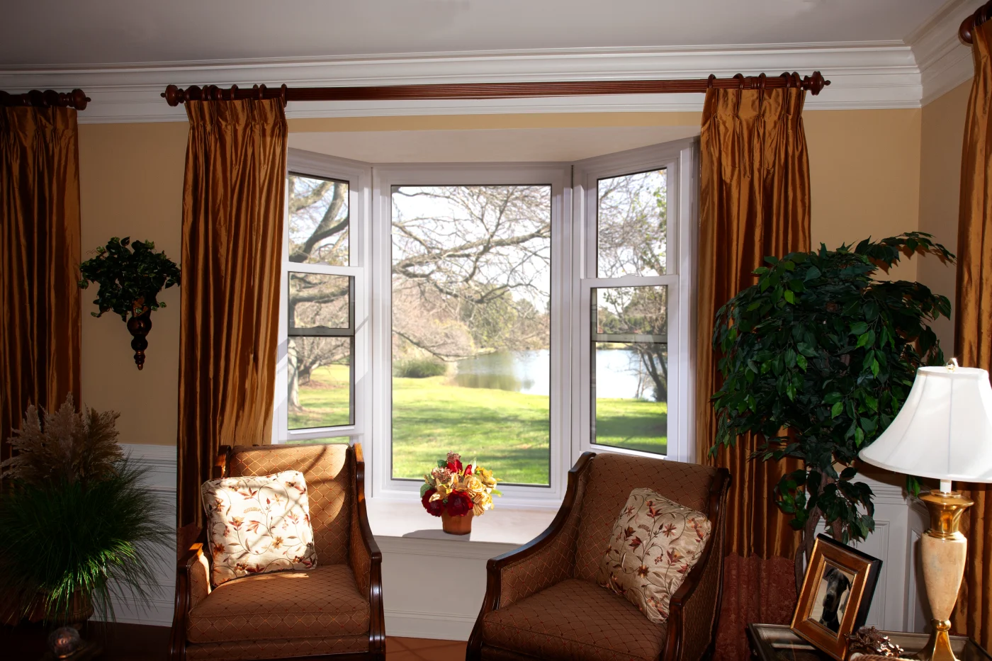 A beautifully installed white OKNA bay window system opens up to a serene lakeside view, flooding the room with natural light. The center window is fixed, flanked by two operable casement windows angled outward. Inside, the space is warm and elegant, with rich golden drapes, classic armchairs, floral pillows, and a potted flower arrangement on the window ledge. Crown molding and detailed woodwork frame the scene, blending traditional charm with modern functionality.