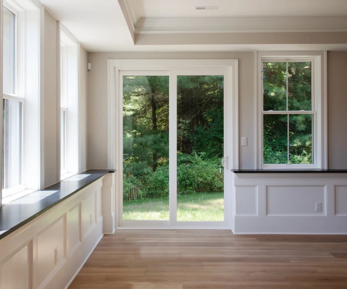 A bright, open living space featuring an OKNA Elegante sliding patio door in a crisp white frame. The wide glass panels create a seamless connection to the lush green landscape outside, while the door’s slim profile allows maximum natural light to flood the room. Clean trim details, soft beige walls, and warm wood flooring highlight the door’s sleek, modern design – perfectly balancing style, security, and energy efficiency for NY Metro homes.