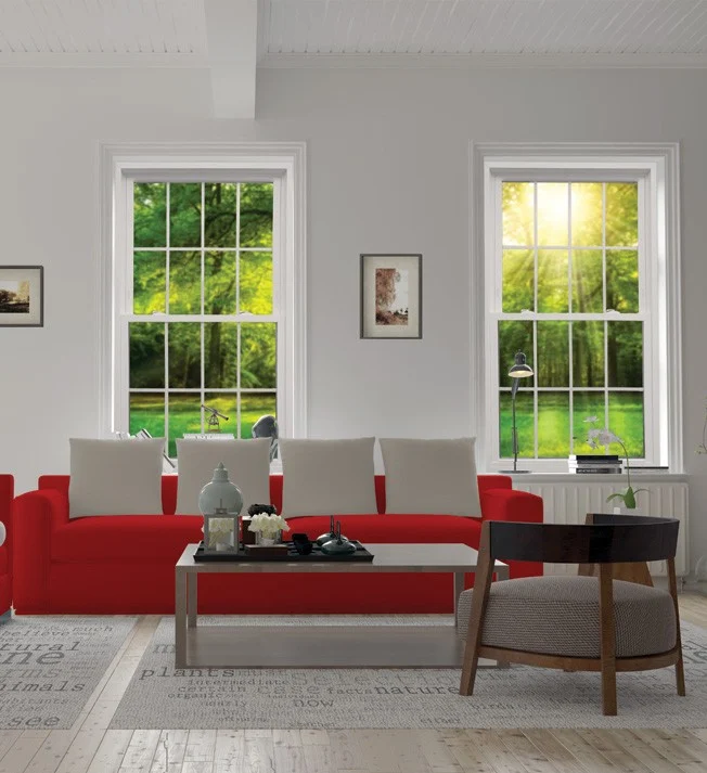 A pair of OKNA double-hung windows—likely from the Precision Weld 400 Series – installed in a bright, modern living room. Sunlight pours through the energy-efficient glass, highlighting the crisp white frames and traditional grille pattern. The windows offer clear views of a lush green yard, emphasizing the beauty, performance, and clean design that OKNA is known for.