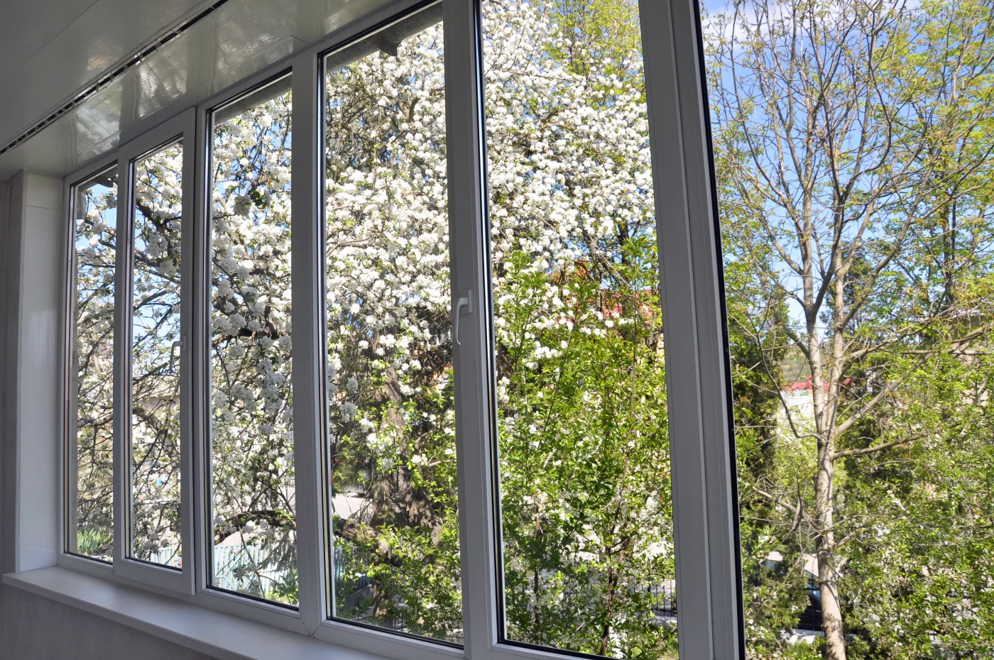A clean, professional window installation featuring a wide, multi-panel design with slim white frames. The windows offer expansive, uninterrupted views of blooming trees and blue sky, emphasizing clarity, craftsmanship, and energy-efficient performance – perfect for brightening up any NY Metro home with natural light and a modern finish.