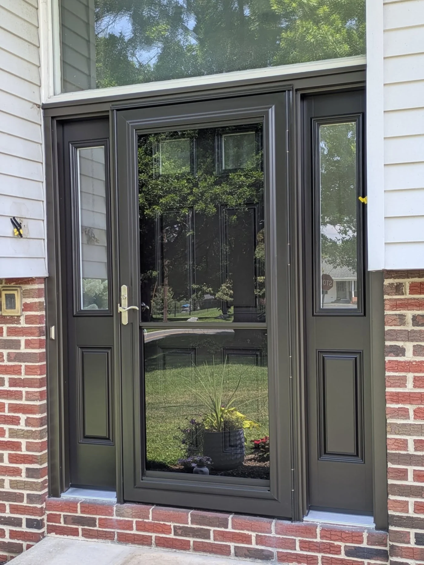 A ProVia aluminum storm door in a deep bronze finish, featuring full-view glass for maximum visibility and light. The door is framed by matching sidelites and set within a brick-and-vinyl exterior, offering superior weather protection for NY Metro homes.