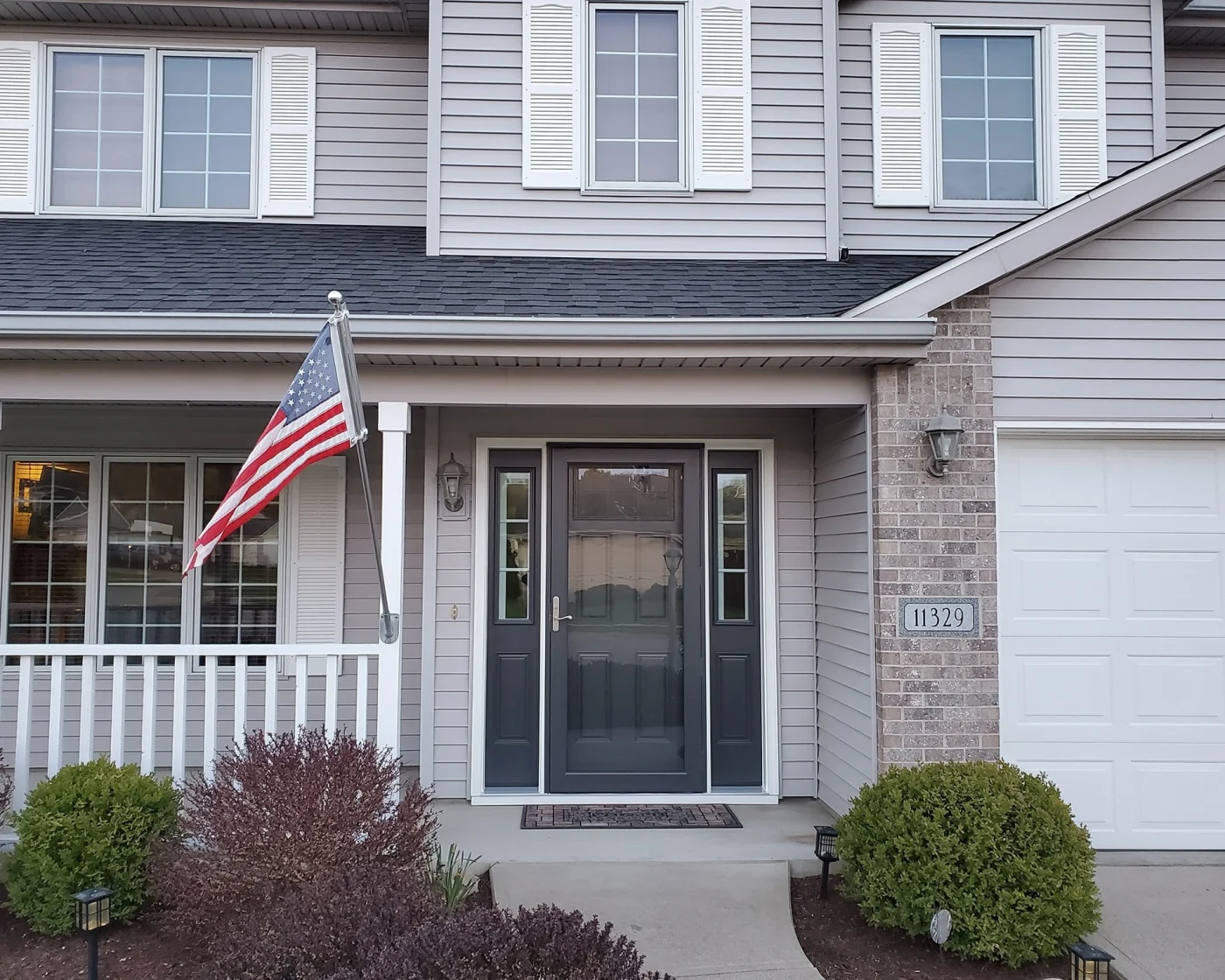 A dark gray ProVia Decorator storm door with full-view glass and matching sidelights, installed on a two-story home with light gray siding, white shutters, and brick accents. An American flag is mounted on the front porch.
