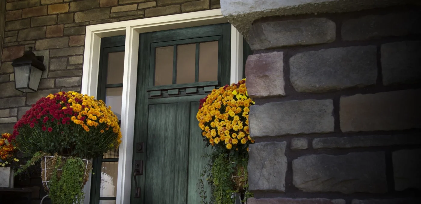 A ProVia Embarq fiberglass front door in a rich, deep green finish, framed by white trim and flanked by tall sidelites. Vibrant red and yellow potted flowers add warmth and charm to the stone-clad entryway – showcasing style and durability for NY Metro homes.