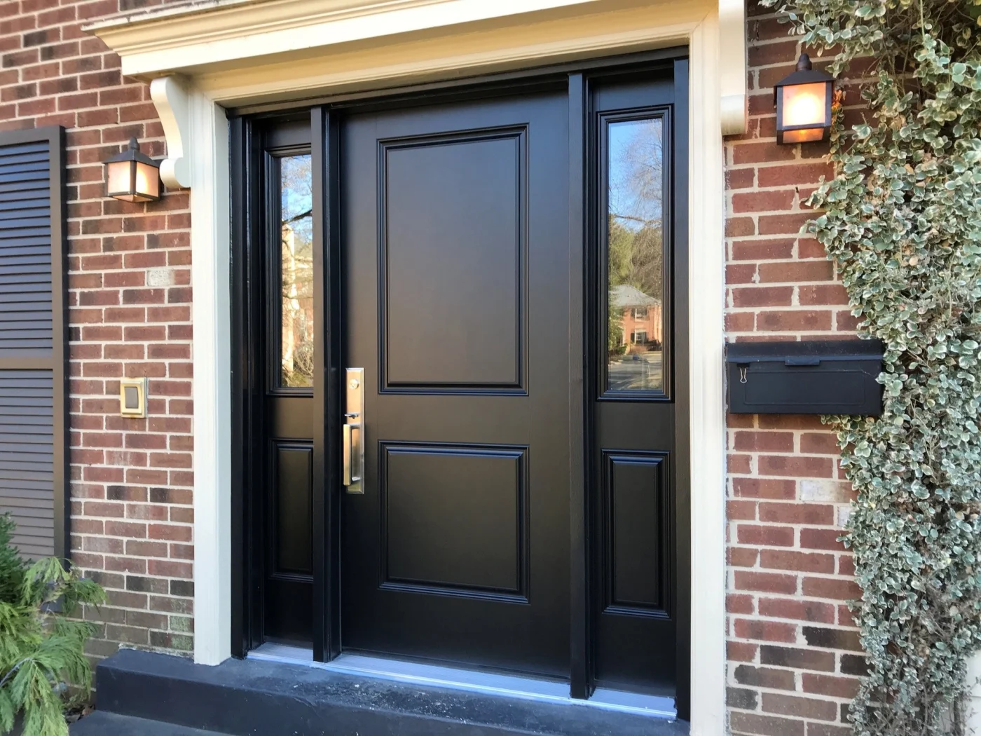 A bold ProVia Heritage fiberglass front door in a deep black finish, framed by matching sidelites with clear glass. The door is set within a red brick exterior accented by cream trim, black shutters, and warm wall-mounted lanterns, creating a stately, upscale NY Metro entryway.