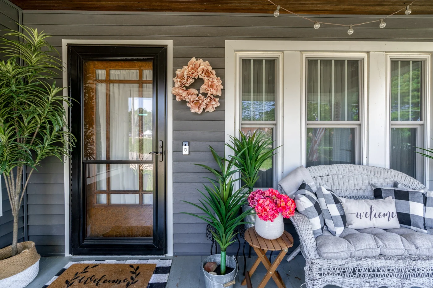 ProVia Storm Door Installation | NY Metro, Tri-State Region | Window King A full-view ProVia Spectrum storm door in a coal black finish, installed on a porch with dark gray siding and white trim. The door features a large glass panel with a woodgrain-look interior frame. The surrounding porch décor includes potted plants, a wicker loveseat with plaid pillows, and a “Welcome” doormat.