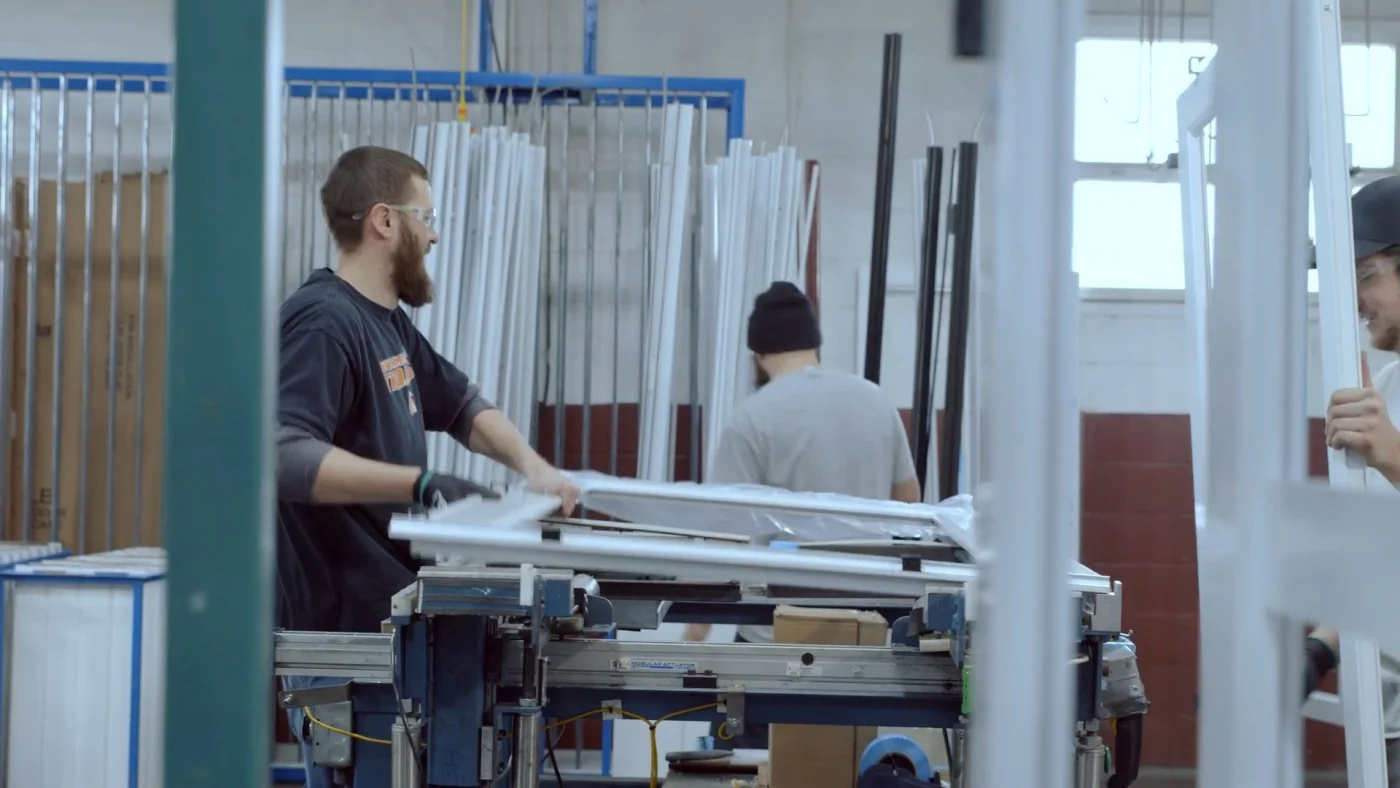 Workers assemble ProVia Spectrum storm doors inside a manufacturing facility. White aluminum frames and components are arranged around the workspace, with production equipment and stacked materials visible in the background.