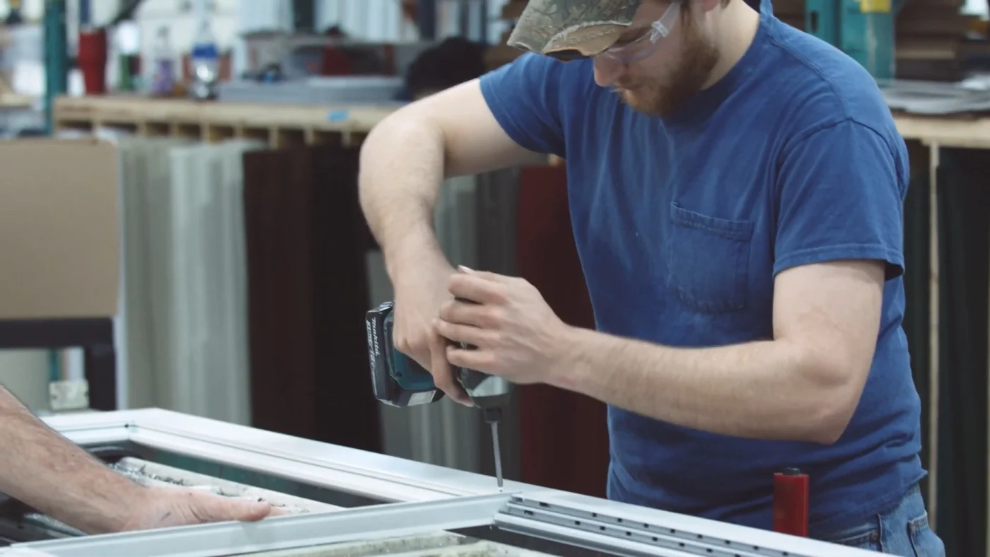 A worker uses a power drill to assemble a ProVia storm door frame inside a manufacturing facility. The door components are laid flat on a work surface, with other tools and materials visible in the background.