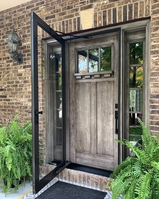 A black ProVia Superview storm door with a full-view glass panel, shown open in front of a rustic wood entry door with matching sidelights. The home features a brick exterior, decorative lighting, and lush potted ferns on either side of the entrance.