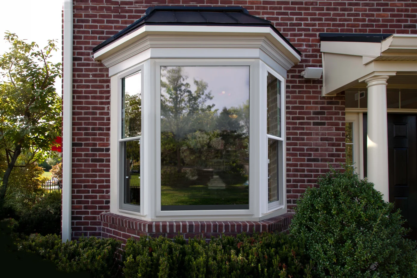 This exterior shot showcases a beautifully installed Starmark Evo Bay window on a traditional red brick home. The three-panel unit features a large center picture window flanked by two operable casement windows, all framed in a clean, white composite material. The window extends outward with a graceful curve, creating both architectural depth and expanded interior space.