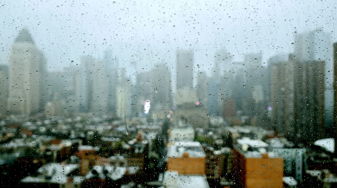A rainy New York cityscape viewed through a window covered in water droplets. The skyline, slightly blurred in the background, suggests an overcast, stormy day, while the raindrops in sharp focus on the glass highlight the barrier between the cozy indoors and the wet, unpredictable weather outside. It visually underscores the importance of durable, weather‑tight windows in handling the New York Metro’s wild seasonal swings.