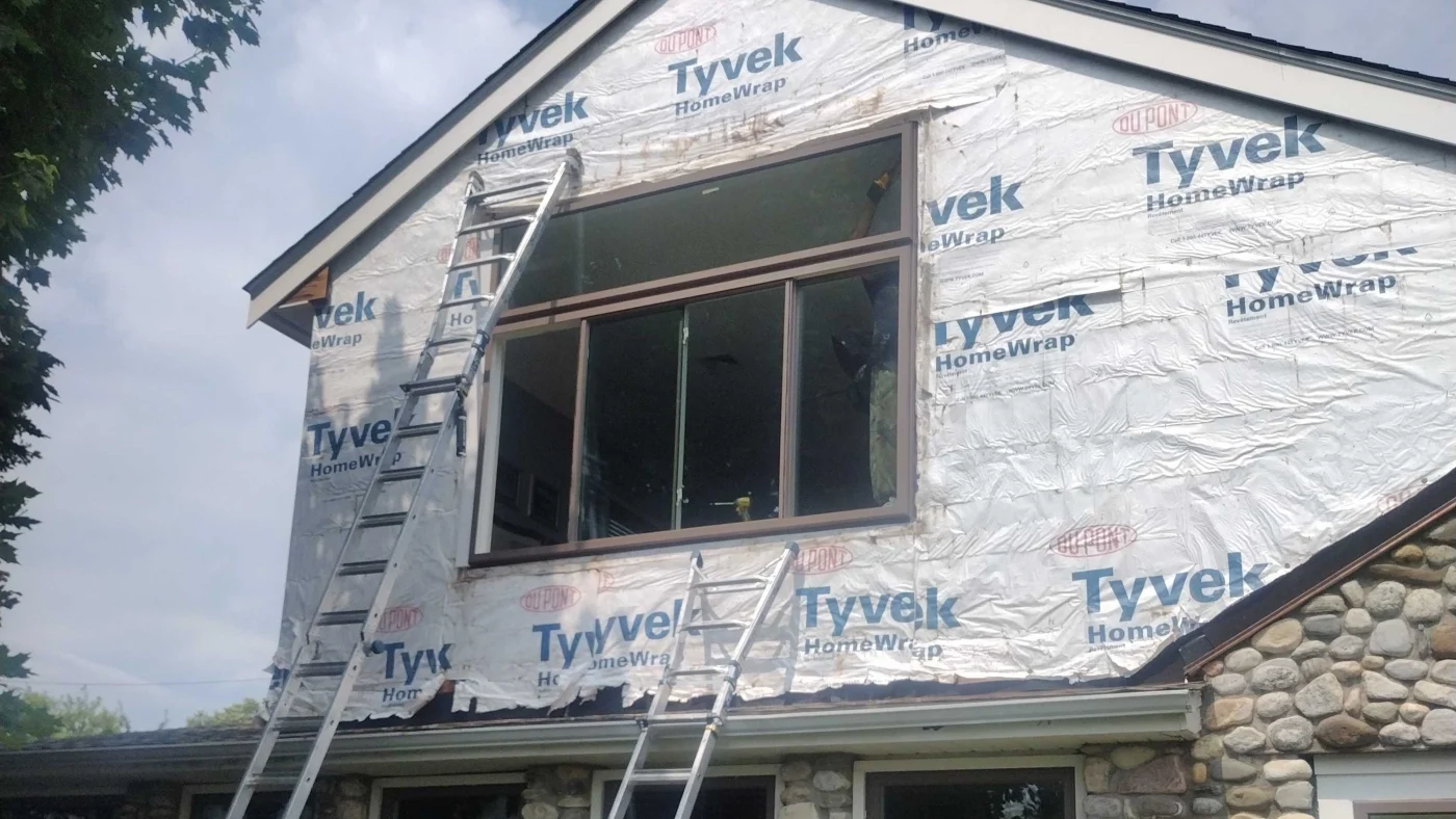 A large window installation in progress on a home, with Tyvek house wrap visible and ladders set against the siding.