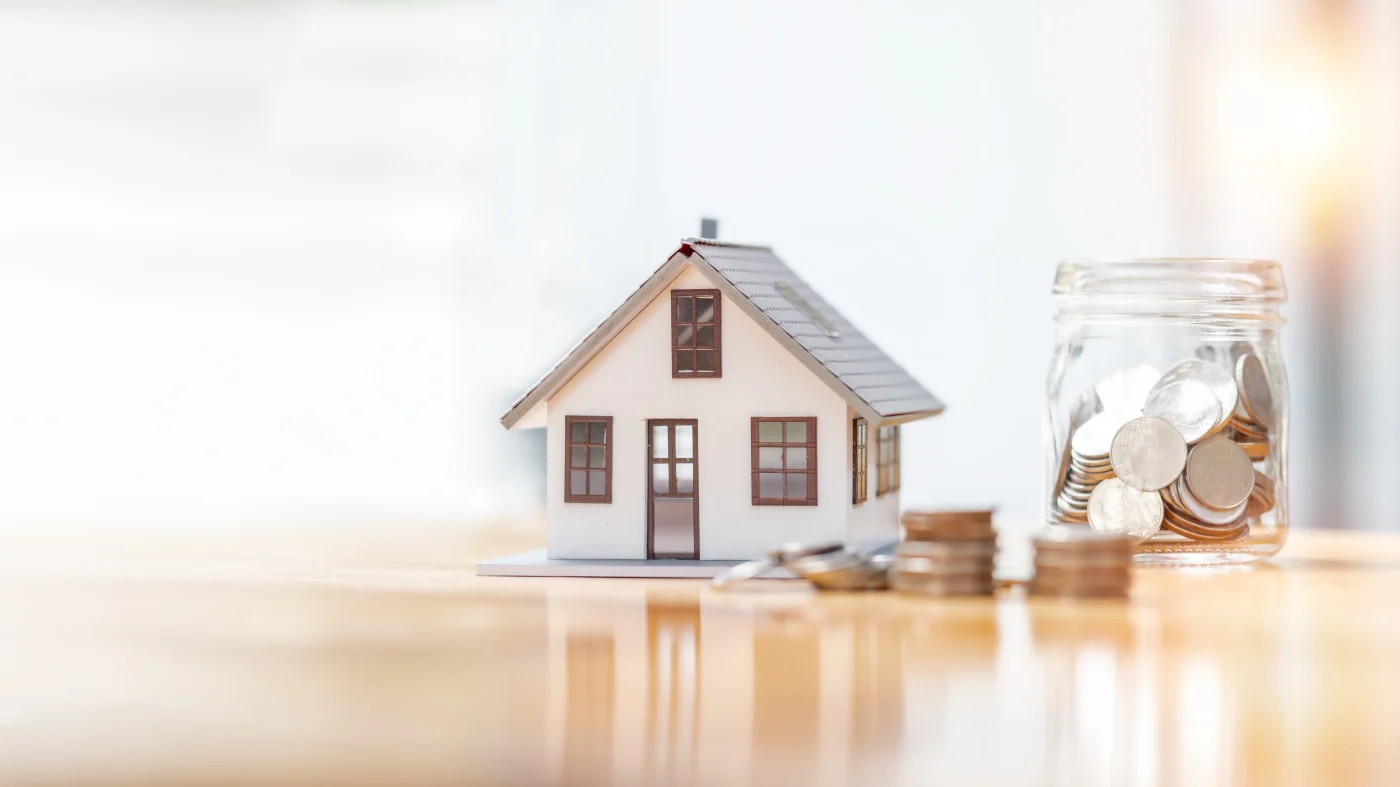 A small glass jar with silver coins in it, along with some coins stacked in front of a small home, symbolizing the growth of property value through proper investment.