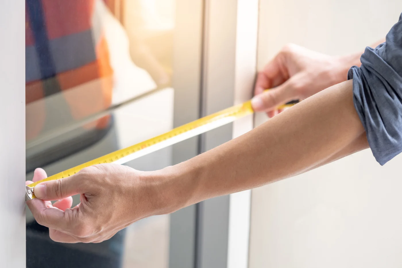 A close-up of a window installer carefully measuring the width of a window frame with a yellow tape measure. Sunlight filters in, highlighting the clean lines of the frame and the installer’s rolled-up sleeves – capturing the precision, professionalism, and hands-on care that define a job done right the first time.