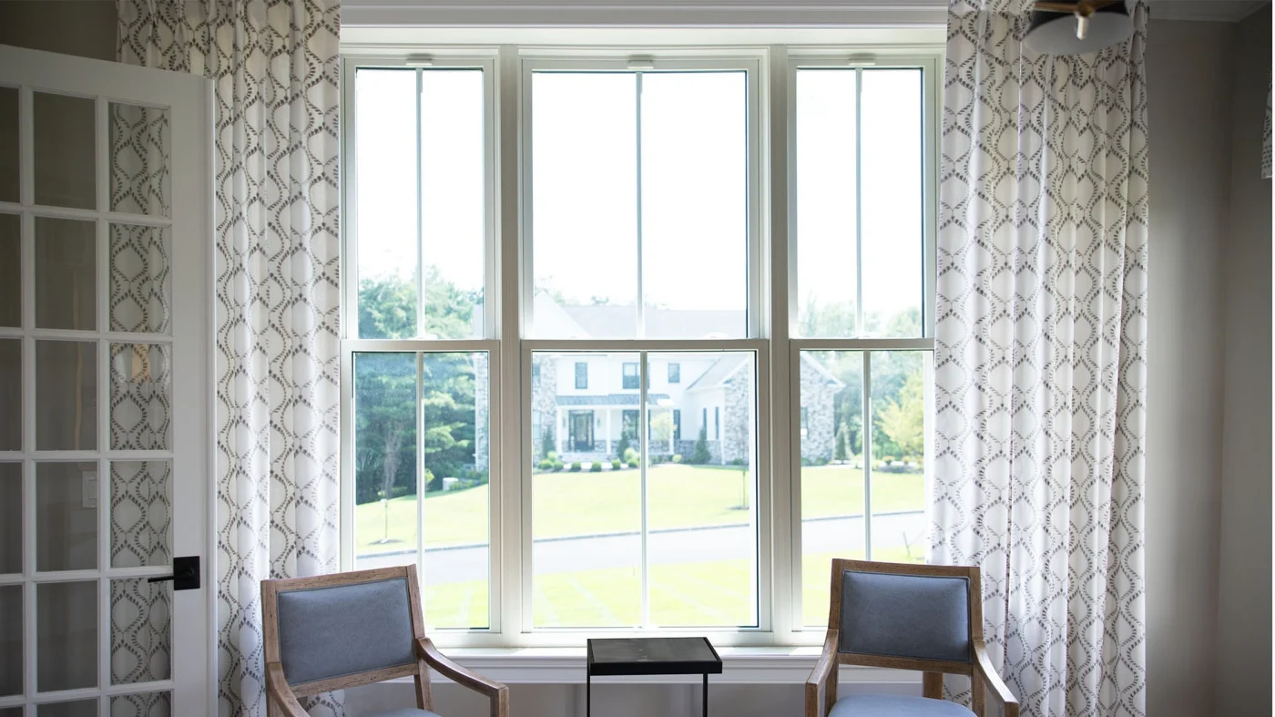 A pristine, sunlit room with tall white-trimmed double-hung windows framed by elegant patterned curtains. The windows offer a crystal-clear view of a well-kept suburban street and home. Everything – from the trim lines to the glass—is flawlessly clean and symmetrical, reinforcing the message: at Window King, details matter. Every angle, every finish, every install – done with care.