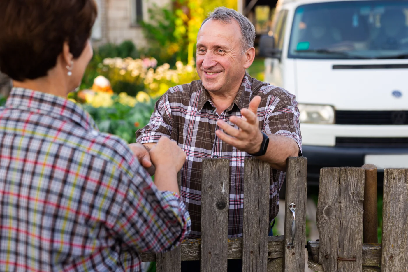 What Sets Window King Apart From NY Metro Window/Door Companies A friendly, genuine moment between two neighbors chatting over a rustic wooden fence. One man is smiling warmly, gesturing with his hands as he talks, while a woman listens attentively. The background features a garden full of greenery and flowers, with a white work van parked nearby—suggesting a recently completed service visit.