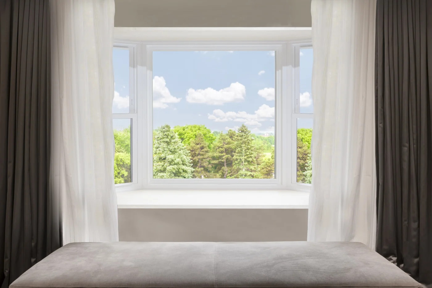 A bright bay window framed by sheer white curtains and darker side drapes, opening up to a beautiful view of lush green trees beneath a blue sky dotted with fluffy clouds. The clean white frame and expansive glass panels allow natural light to flood the room, creating an airy, open atmosphere. The built‑in bench seat beneath the window offers a cozy spot to relax and enjoy the scenery, making it both a functional and elegant architectural feature.