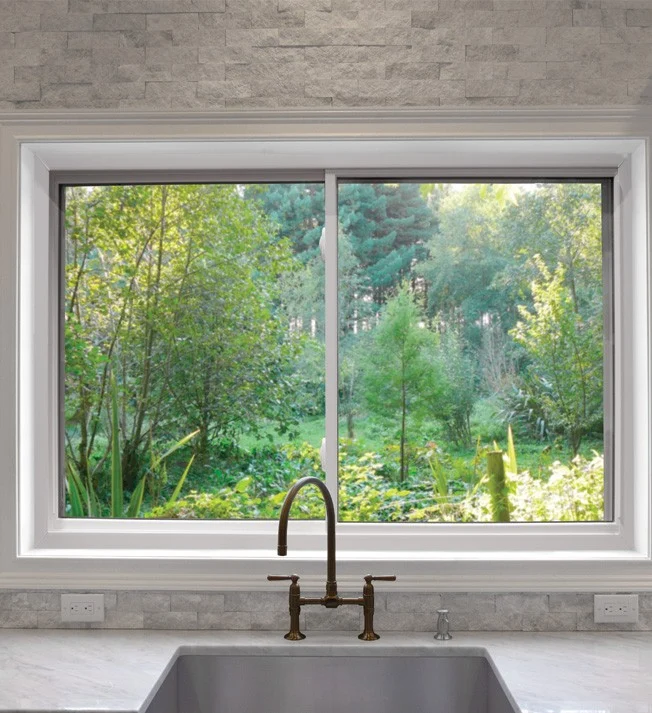 A large OKNA window installed above a kitchen sink, framed by a clean white casing. The sleek, modern window design provides a crystal‑clear view of a lush green backyard filled with trees and shrubs, letting in abundant natural light. The stone‑textured backsplash and brushed bronze faucet add a touch of elegance, while the wide glass panels highlight the openness and connection between the indoor space and the outdoors.