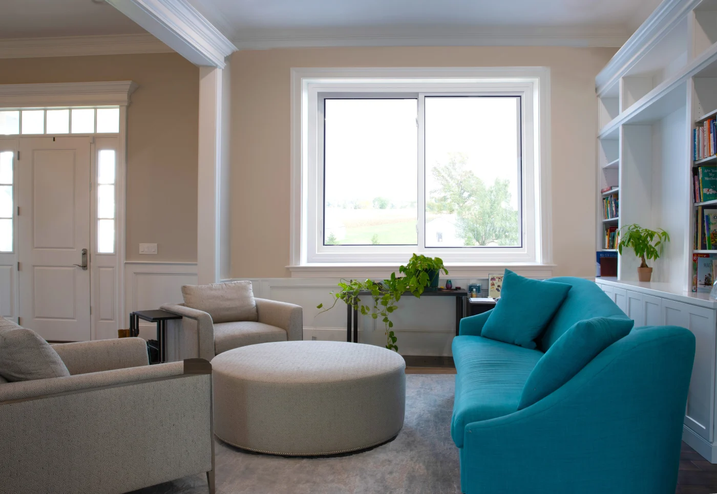 A bright, inviting living room featuring a large, modern sliding window from either the Starmark Evo line. The window’s clean white frame contrasts with the soft beige walls and allows ample natural light to flood the space. Comfortable seating, including a bold teal sofa and neutral armchairs, creates a cozy yet stylish atmosphere. Built‑in bookshelves, potted plants, and tasteful décor complete the look, while the expansive glass offers a clear view of the outdoors.