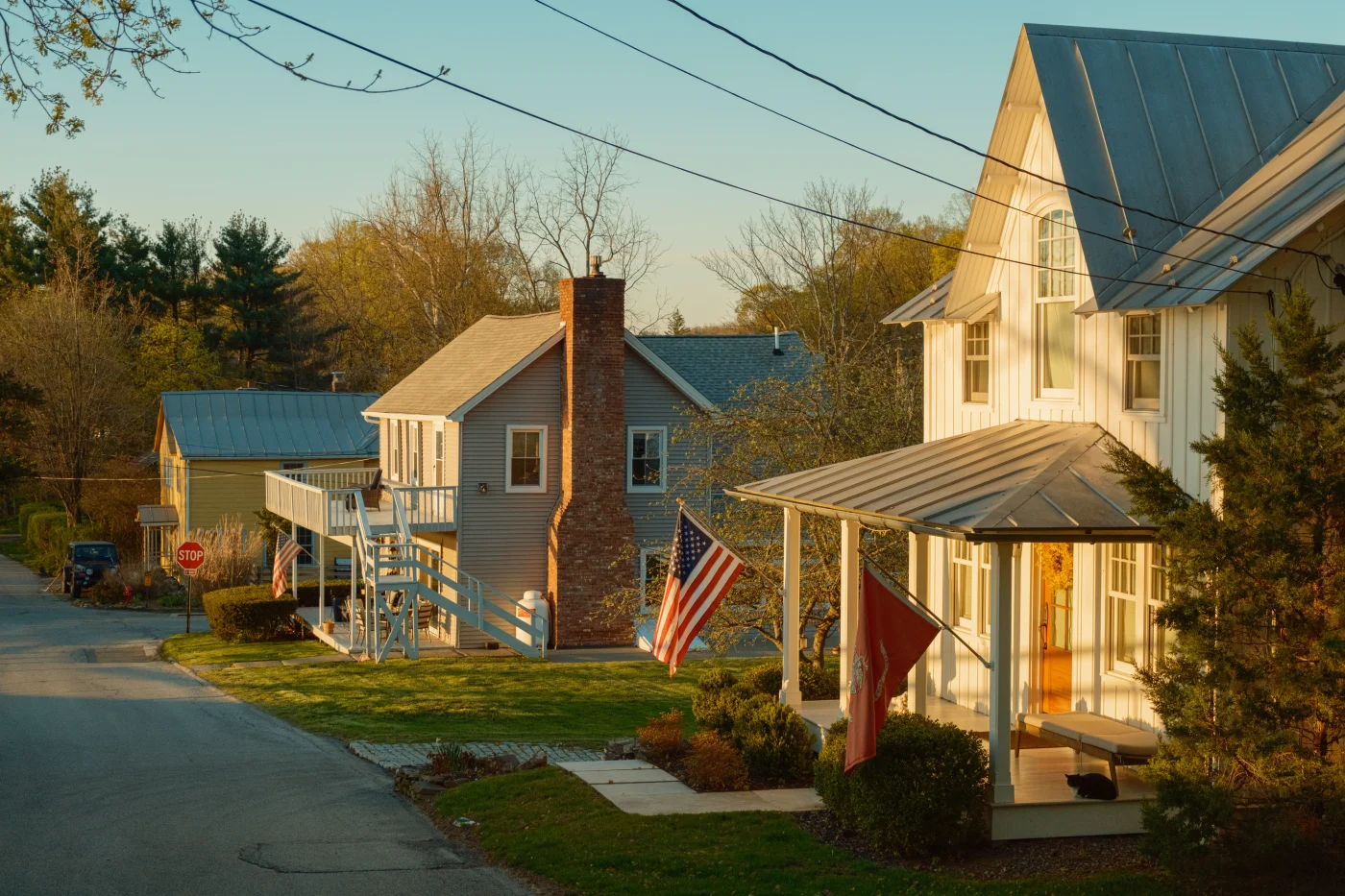 A quiet residential street in the New York area during golden hour, with warm sunlight casting a soft glow over the homes. In the foreground, a white two‑story house with a metal roof and an inviting front porch displays a red flag, while further down the street, other houses—one with light gray siding and a prominent brick chimney—feature American flags.