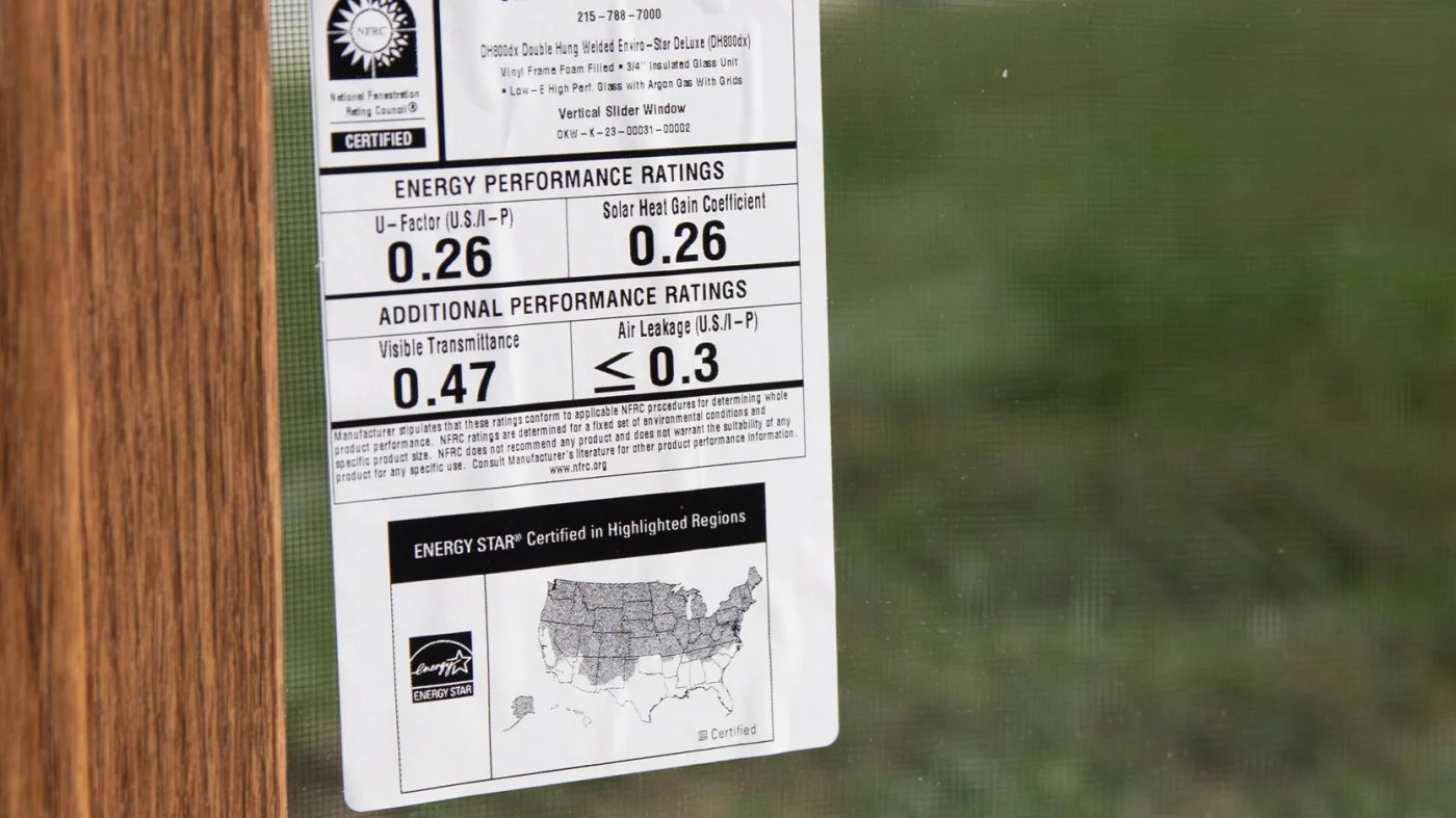 A close-up of the NFRC energy performance label affixed to an OKNA Enviro-Star Series 800 window. The label displays key efficiency metrics, including a U-Factor and Solar Heat Gain Coefficient, and an Air Leakage rating. The background reveals a natural outdoor view through the window, emphasizing clarity and insulation. It’s a clear, data-backed testament to the superior energy efficiency standards Window King insists on for NY Metro homes.