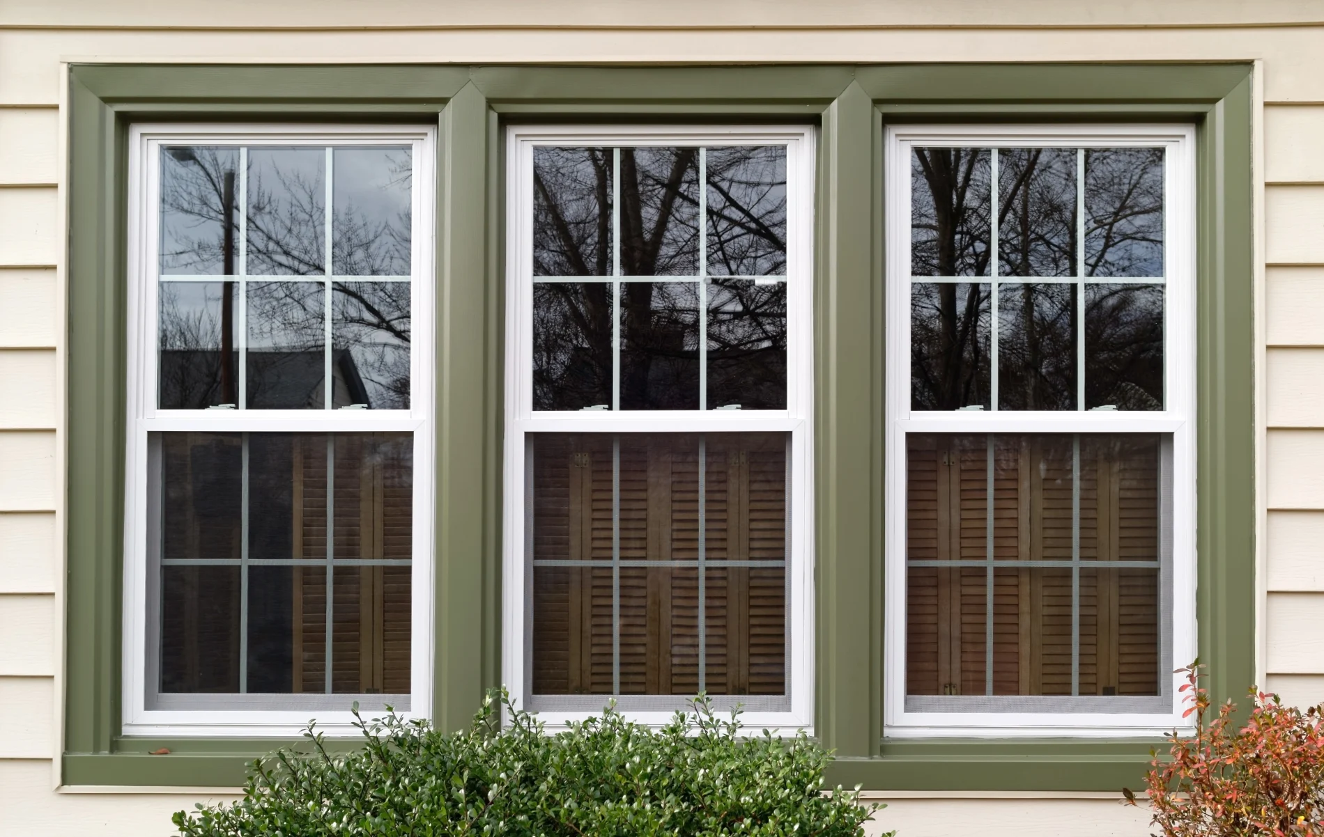 A set of three white double-hung replacement windows with decorative grids, framed in olive green trim, was installed on a cream-colored siding home in Yonkers, NY. The clean lines and symmetrical design highlight professional craftsmanship and energy-efficient materials.