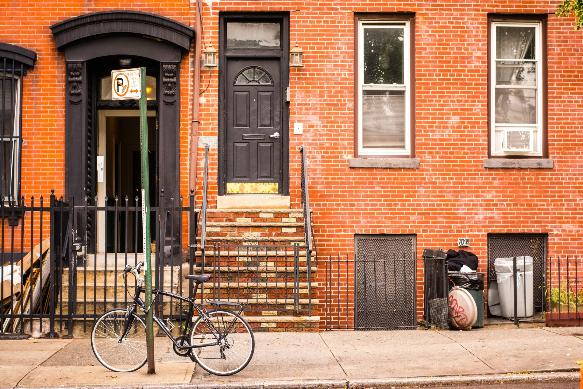 A classic Brooklyn, NY, streetscape featuring a red-brick rowhouse with black trim, a stoop, and tall windows. A bicycle leans casually against a street sign out front, adding to the everyday energy and neighborhood charm that make Brooklyn, NY, blocks so distinctive.