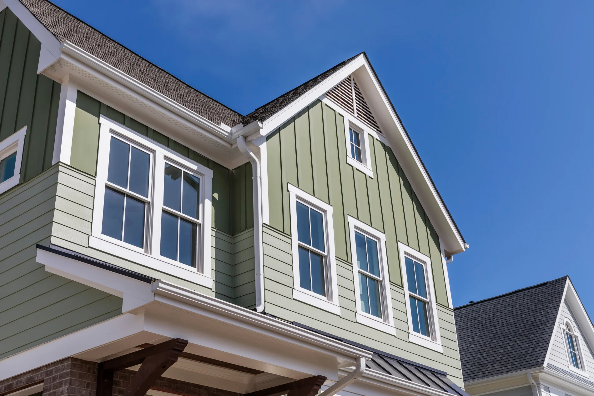 Close-up view of a modern two-story home with green board-and-batten siding, crisp white trim, and multiple energy-efficient windows reflecting a bright blue sky.