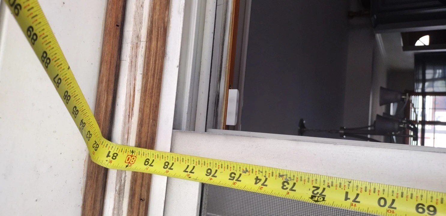 A close-up view of a window measurement in progress, showing a yellow tape measure stretched across a wooden window frame.
