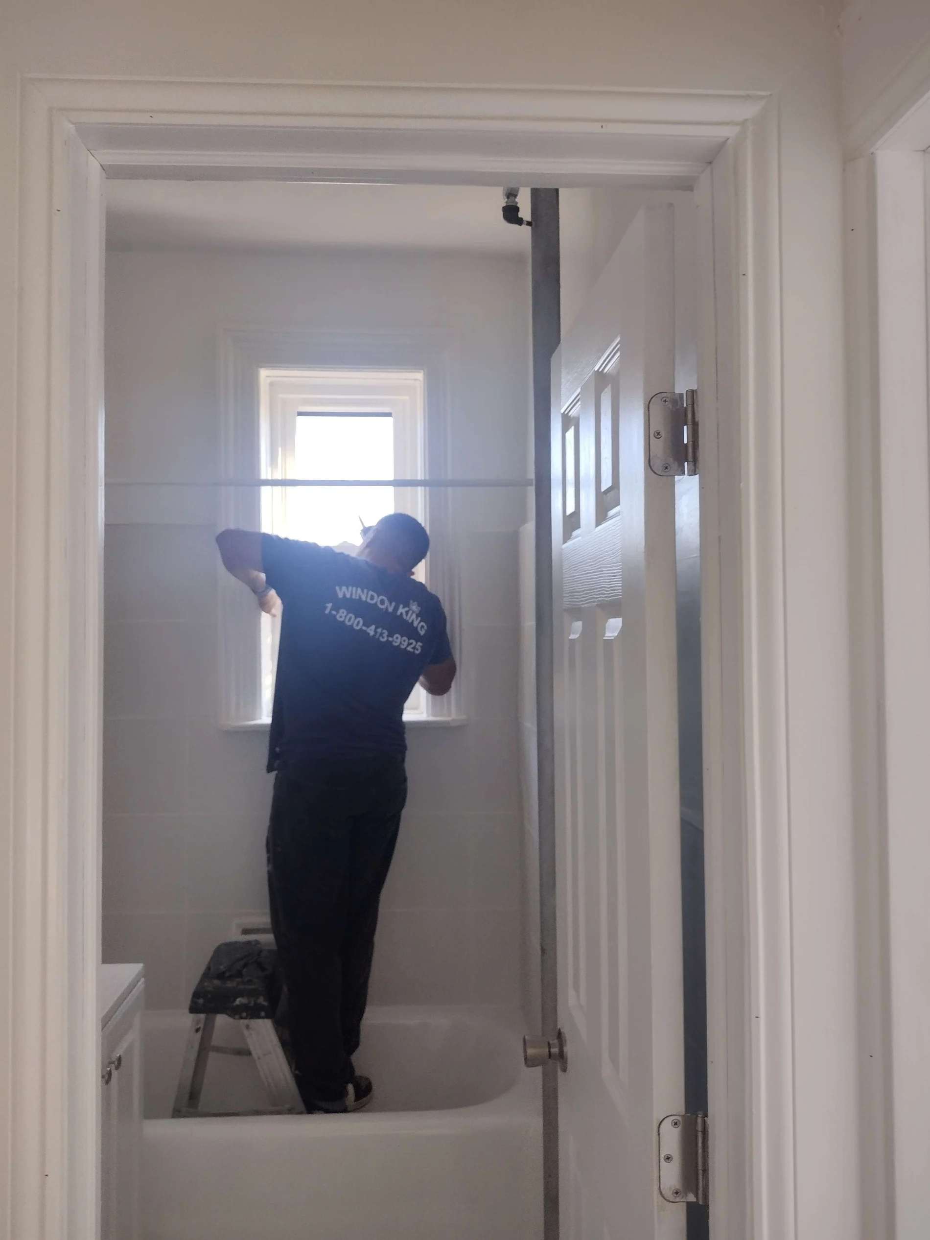 A window installer from Window King stands inside a small bathroom, positioned on a step stool in the bathtub, while working on a narrow window.