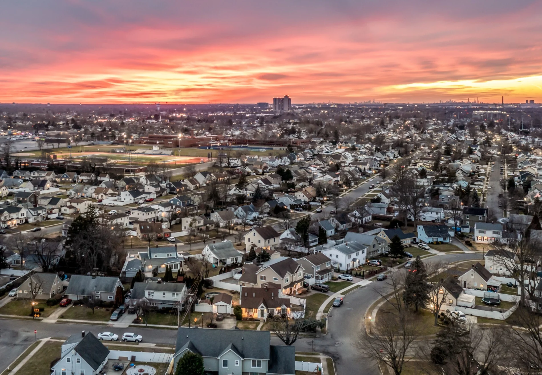 A sweeping aerial view of a suburban neighborhood in Nassau County, Long Island, captured at sunset. Rows of homes with driveways and tree-lined streets stretch across the frame, with a brightly lit sports field visible near the center. The mix of calm suburban streets and the vibrant sunset creates a striking view of everyday life in the NY Metro