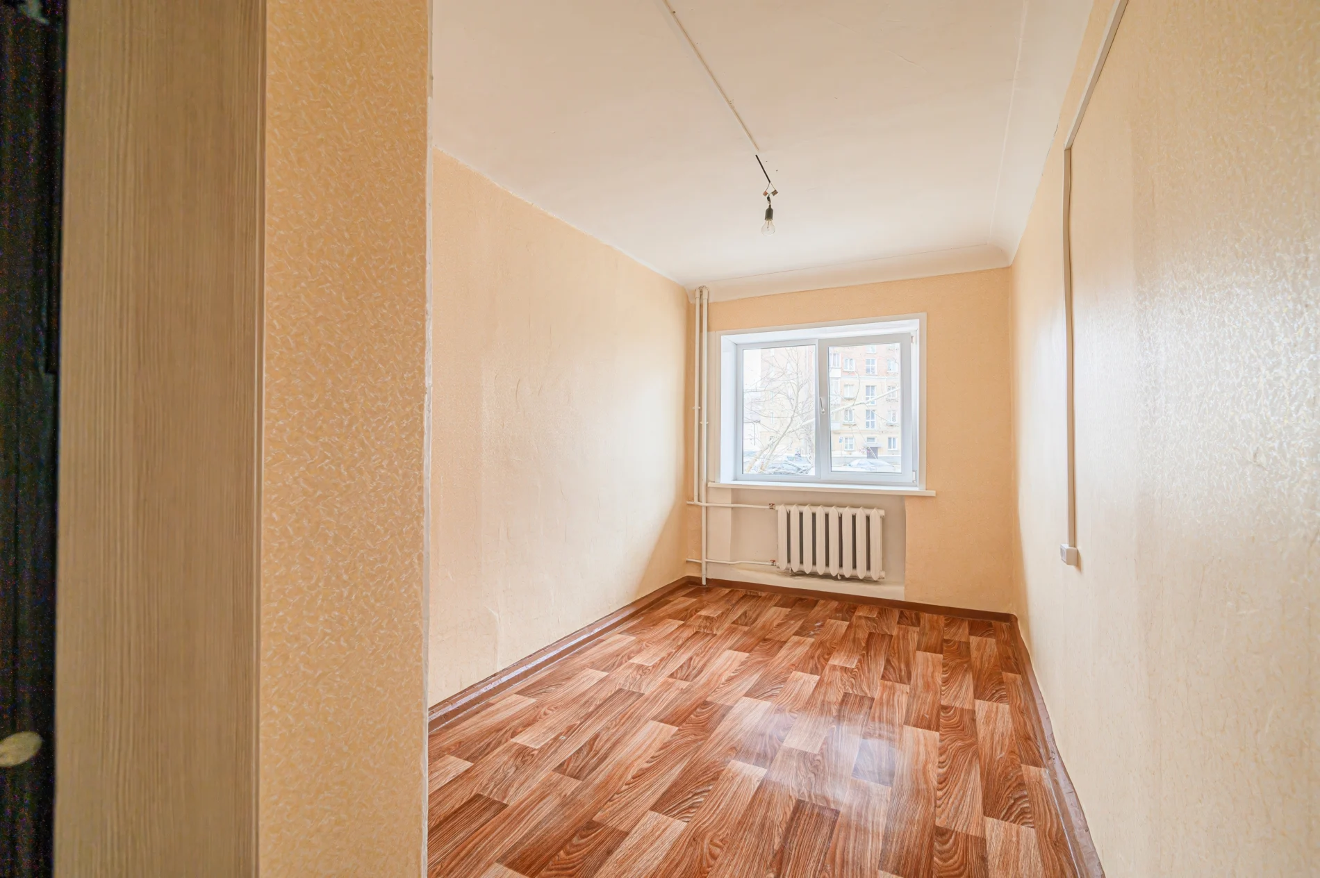 Window Replacement Bronx Ny Affordable Home Windows Window King Interior view of a small, unfurnished room with peach-colored walls, wood-patterned flooring, and a single window overlooking nearby brick apartment buildings. A radiator sits below the window, highlighting the urban residential setting typical of Bronx, NY homes.