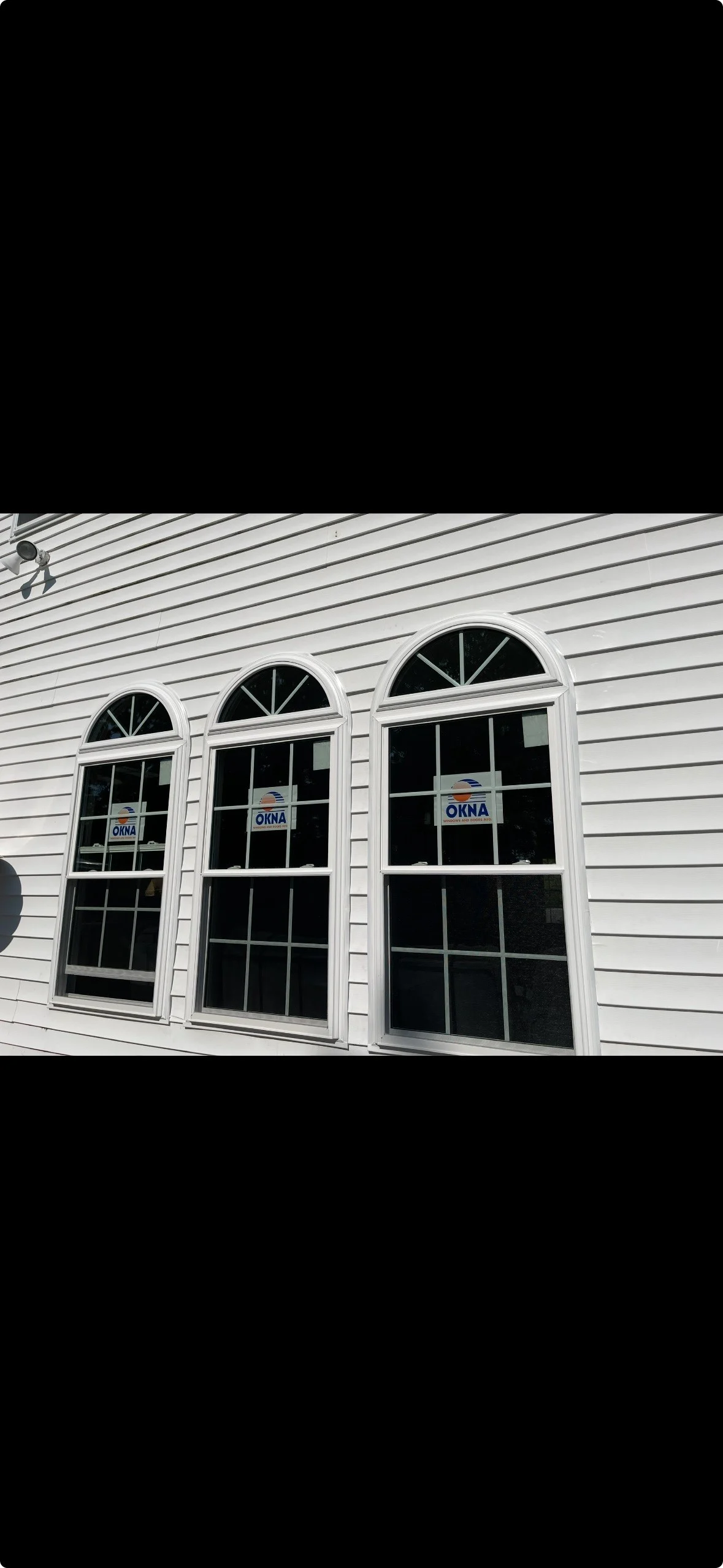 Three white OKNA replacement windows with arched tops installed on a white vinyl-sided house, reflecting sunlight on a bright day.