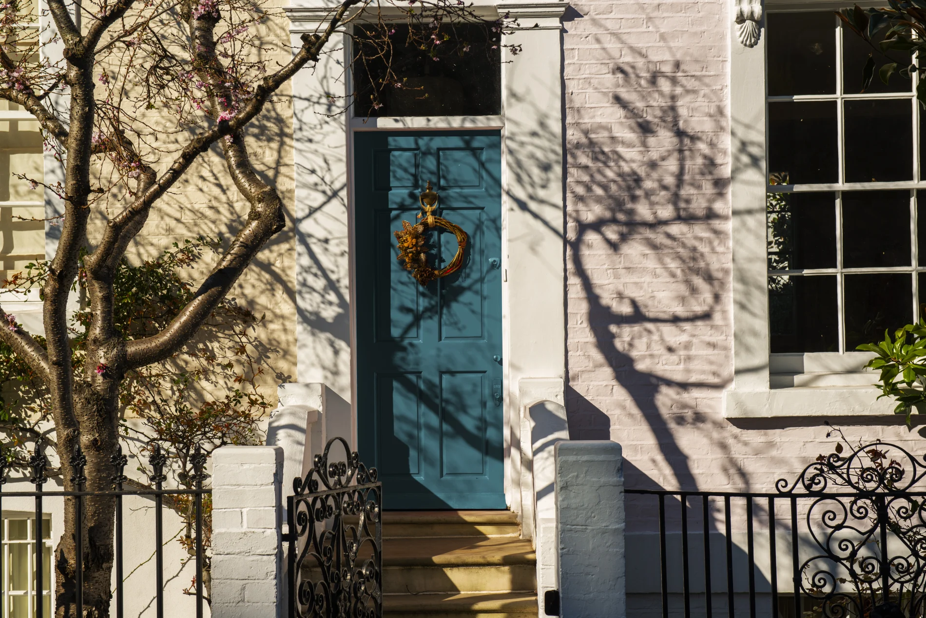 Charming townhouse entry with teal front door, wreath, white brick façade, wrought iron gate, flowering tree, and dramatic sunlight shadows.