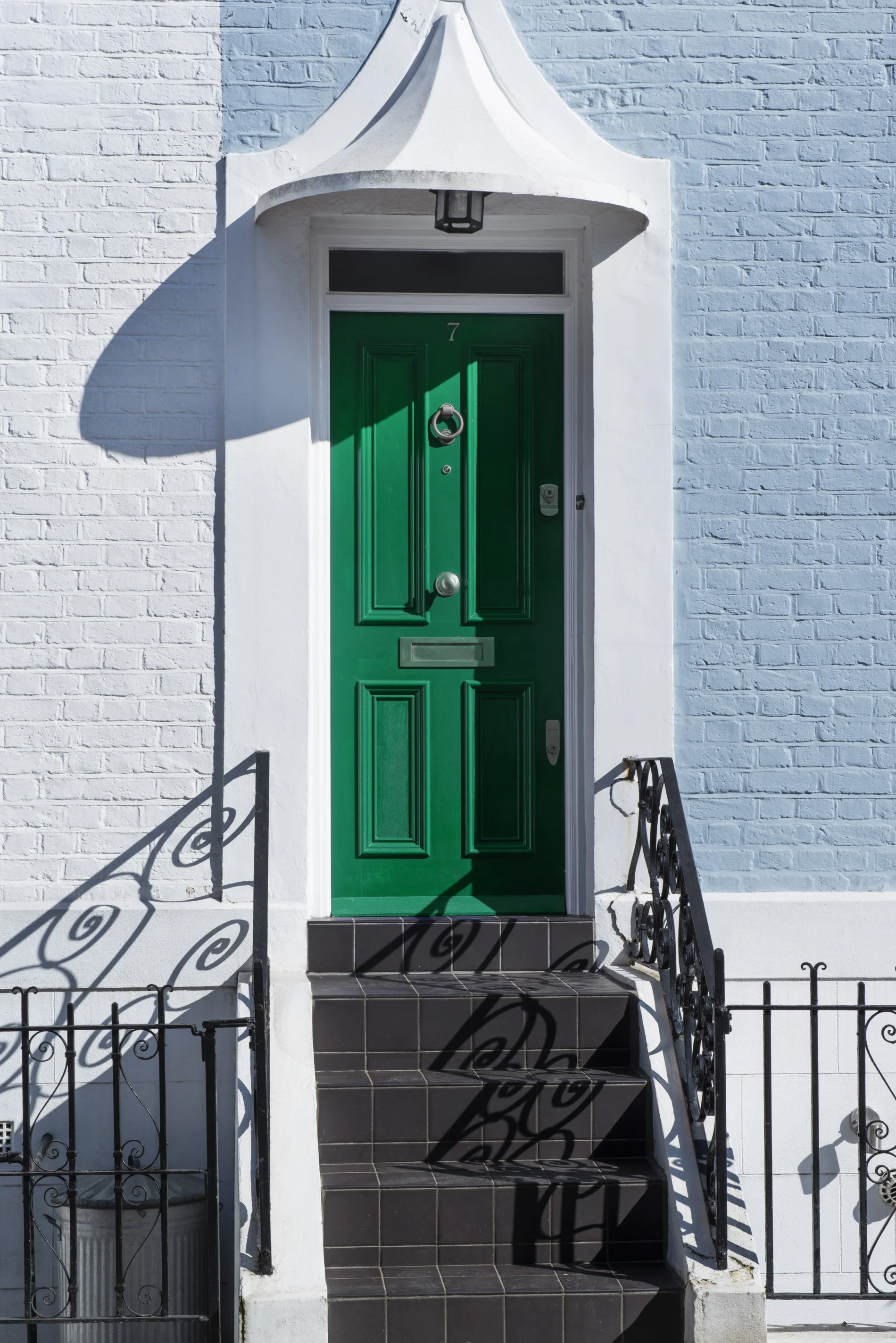 Green front door with black tiled steps, white brick exterior, arched entryway, and iron railings casting crisp shadows in sunlight.