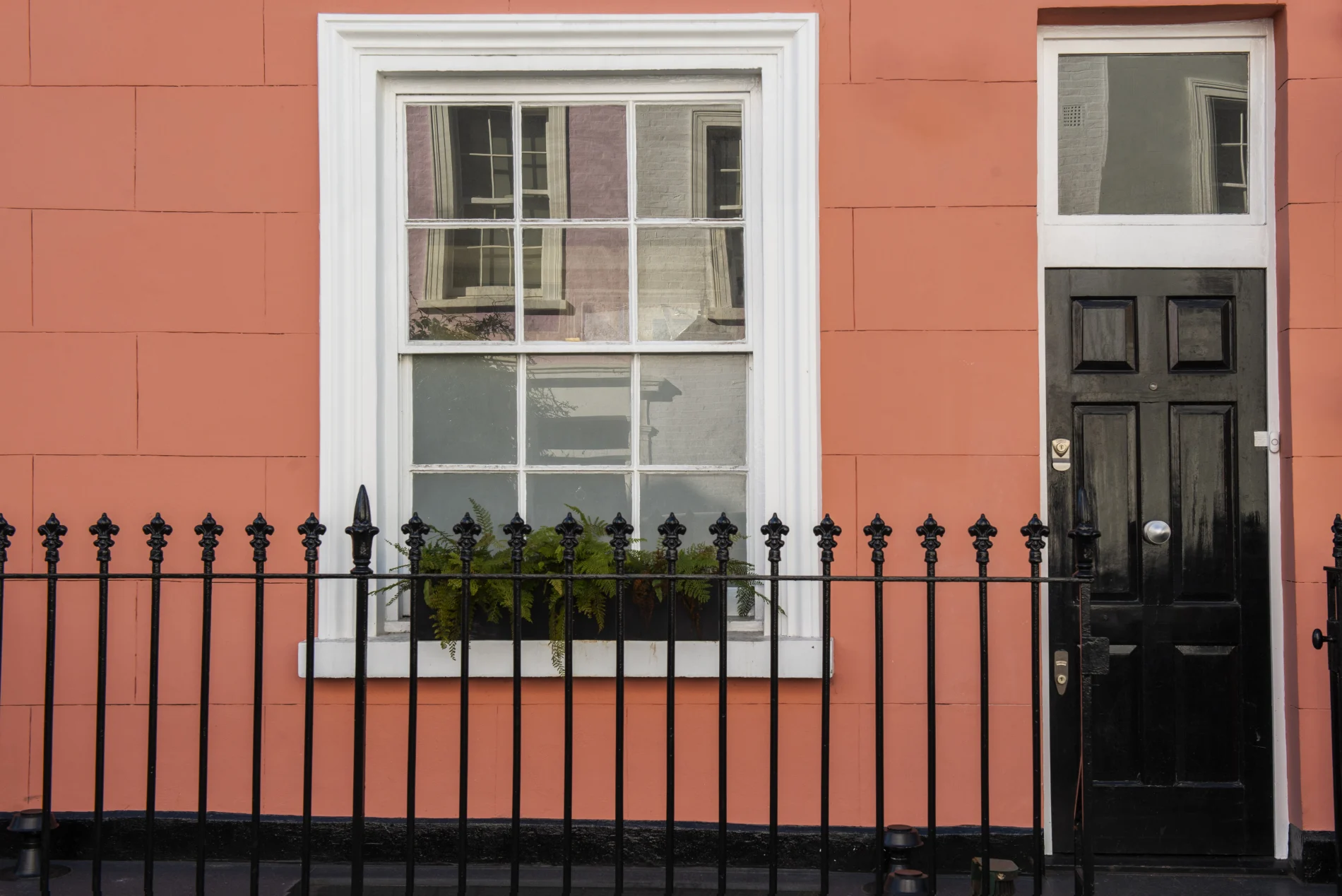 Charming urban townhouse with coral façade, black front door, white-trimmed sash window, wrought iron fence, and window planter with green ferns.