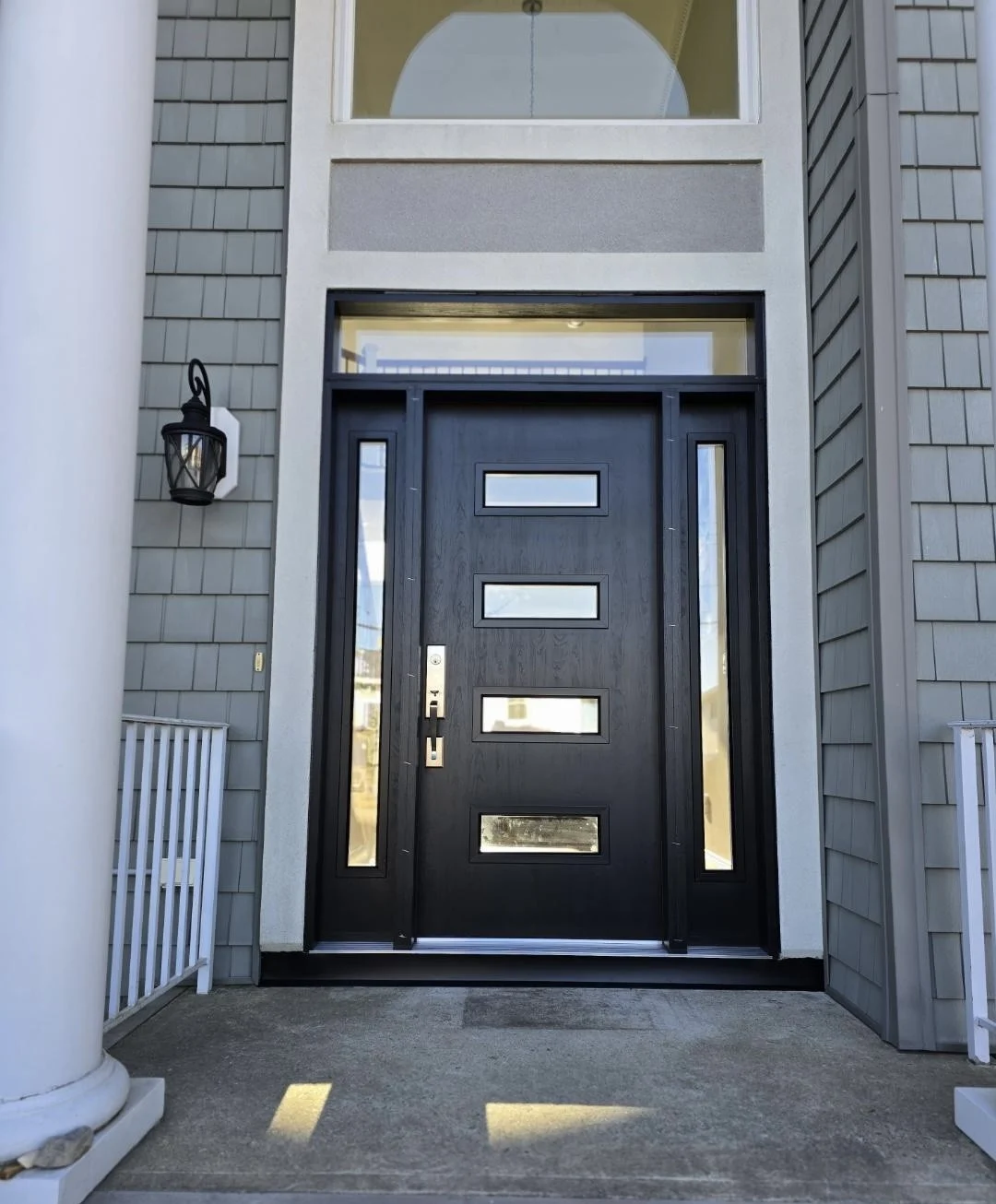 Modern black front entry door with four horizontal glass inserts, narrow sidelights, transom window, gray siding, white columns, and exterior porch lighting.