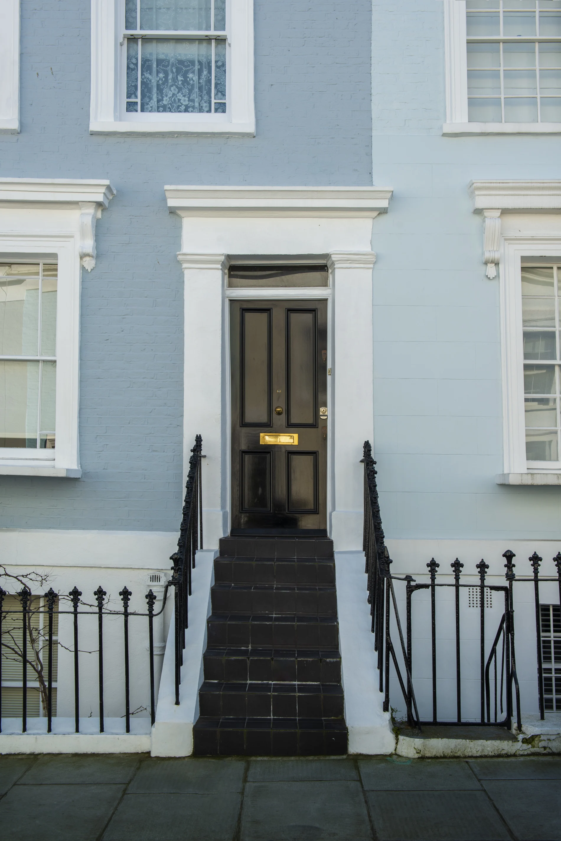 Elegant urban townhouse entry with a black front door, brass mail slot, pale blue façade, white trim, black tiled steps, and wrought iron railings.