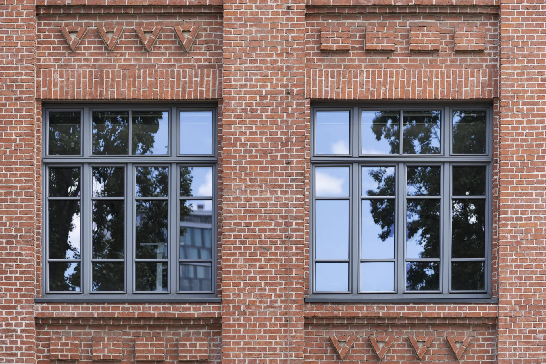 Modern black-framed windows set in a decorative red brick façade, reflecting trees and sky, with symmetrical architectural detailing and urban character.
