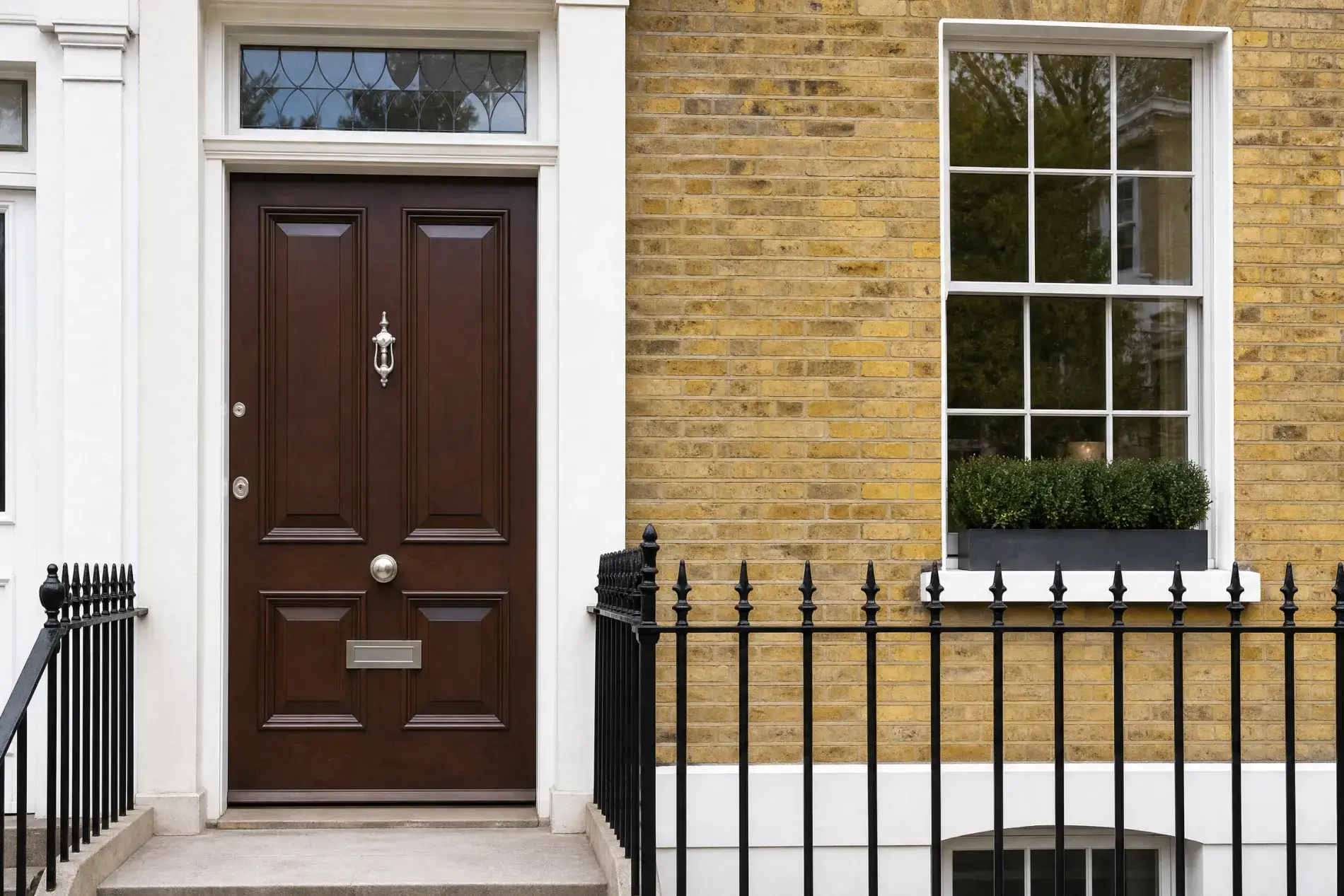 Affordable Door Sales | New Rochelle, NY | Window King Brown paneled front door with silver hardware and a transom beside a yellow brick wall, sash window, and black iron railings.