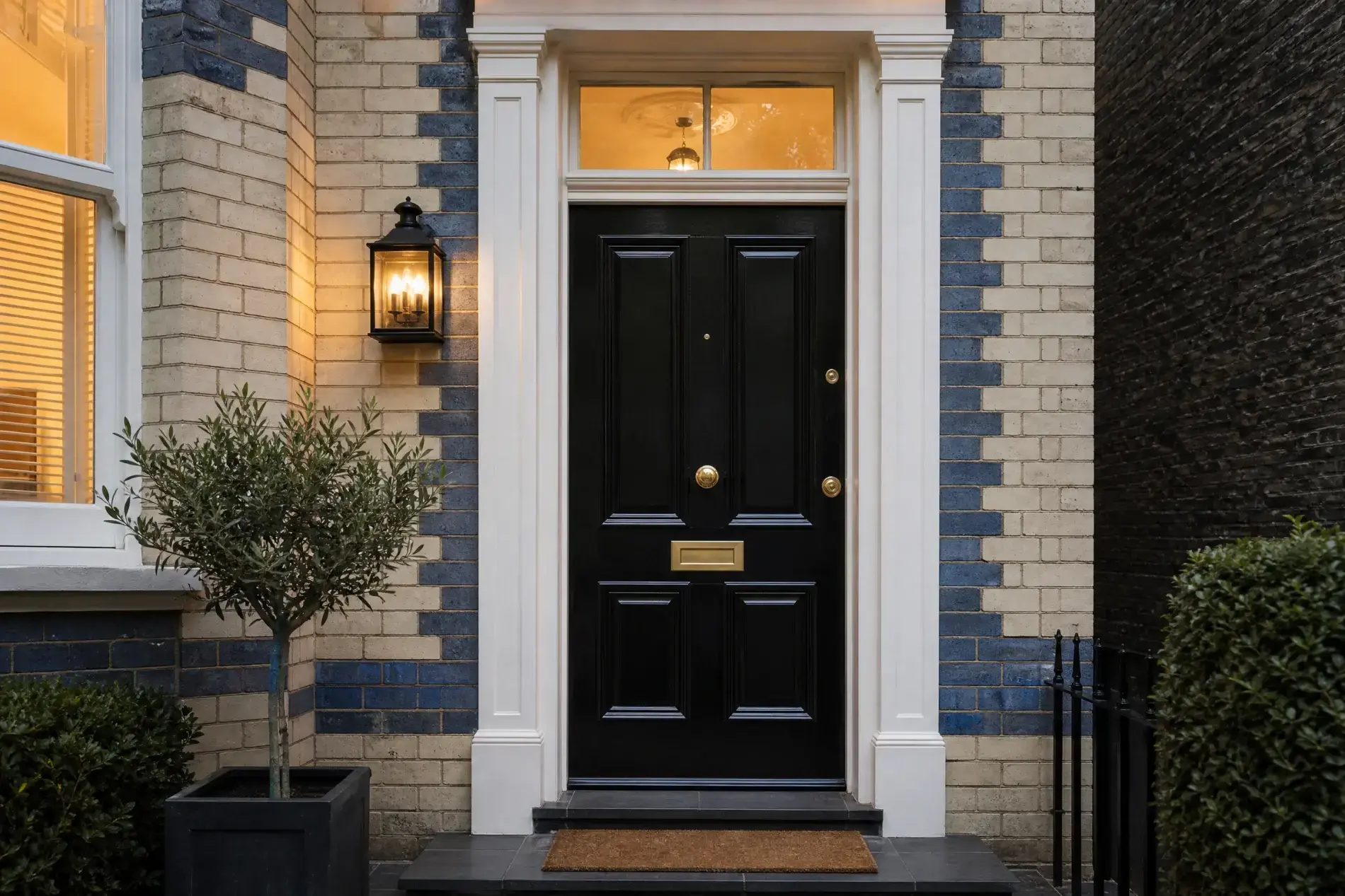 Black paneled front door with brass mail slot and knob, framed in white beneath a transom on a cream-and-blue brick townhouse.