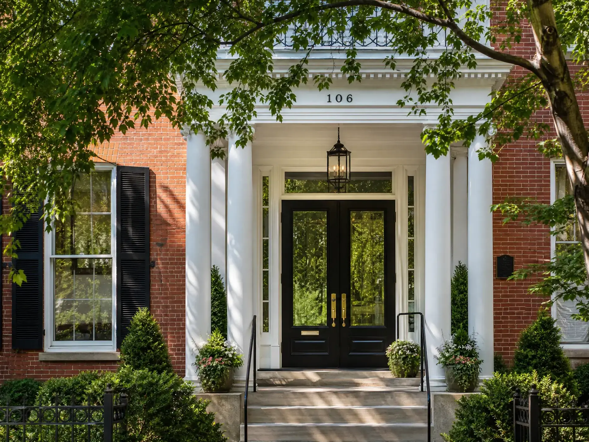 Magnificent Door Installation | Scarsdale, NY | Window King Black double front doors with tall glass panels beneath a white columned portico on a red brick townhouse shaded by leafy branches.